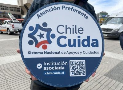 "Un cartel redondo con el texto 'Atención Preferente' en la parte superior, acompañado del logotipo de 'Chile Cuida', que incluye figuras estilizadas de personas en círculo con una estrella en el centro y los colores azul y rojo. Debajo, se lee: 'Sistema Nacional de Apoyos y Cuidados'. Más abajo, un texto indica: 'Institución asociada', junto con un código QR y la dirección web 'chilecuida.cl'. El cartel está sostenido por una persona en un espacio público con vehículos y edificios de fondo."