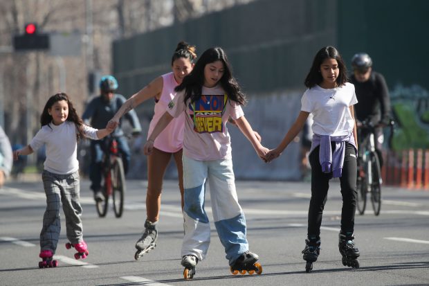 Un grupo de cuatro niñas y una mujer patinan por una calle despejada y cerrada al tránsito vehicular. Tres de ellas usan patines en línea y una lleva patines de cuatro ruedas. Van tomadas de la mano, mostrando equilibrio y concentración mientras avanzan. Al fondo, ciclistas también disfrutan del espacio. La escena transmite una sensación de diversión y actividad al aire libre en un entorno urbano.