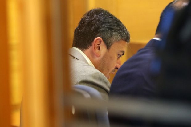 Diputado Ojeda vestido de traje sentado en un tribunal, con expresión seria, observado desde detrás de una barrera.