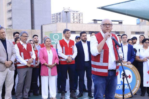 Eric Campos, de chaleco rojo, habla en un micrófono durante un evento al aire libre, rodeado de personas con vestimenta institucional y trajes formales. De fondo, se ven edificios.