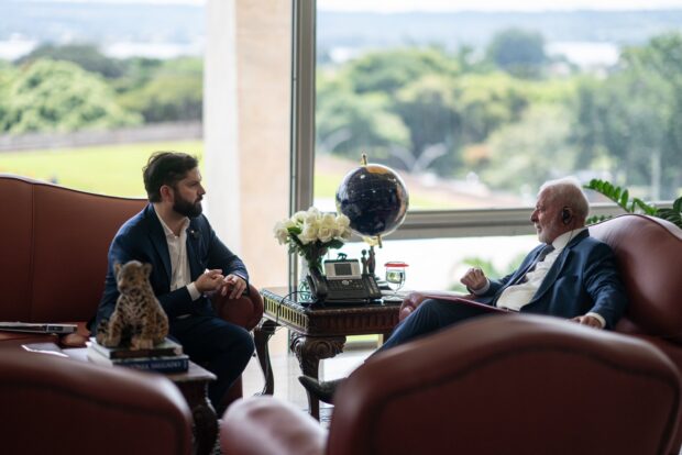Boric y Lula sentados frente a frente en dos sillones distintos, en el medio una mesa con un globo terraqueo. Al fondo un ventanal por donde se ven arboles.