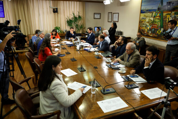 Alrededor de 10 parlamentarios y parlamentarias sentadas en una mesa rectangular con papeles en la mesa.