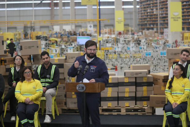 Presidente Boric da un discurso y atrás se múltiples cajas con la cinta amarilla de Mercado Libre