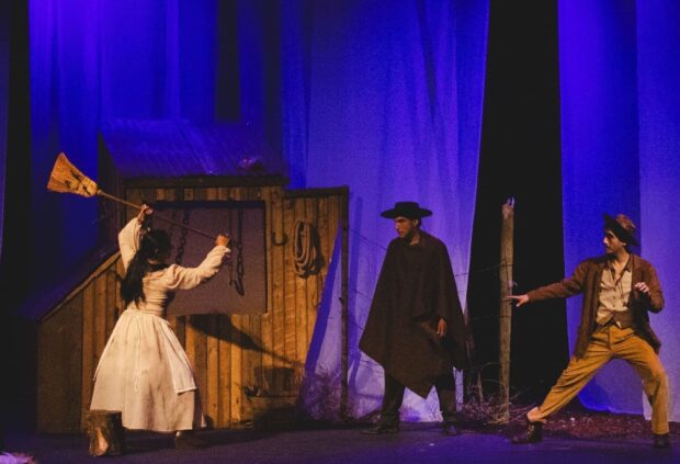 Escena teatral con tres actores en el escenario iluminado con luces azules. Una mujer vestida de blanco levanta un objeto similar a una escoba, mientras dos hombres con atuendos de época, uno con capa y sombrero y otro con chaqueta y pantalón claro, adoptan posturas dramáticas. El fondo tiene un telón con efectos de iluminación que generan sombras alargadas.