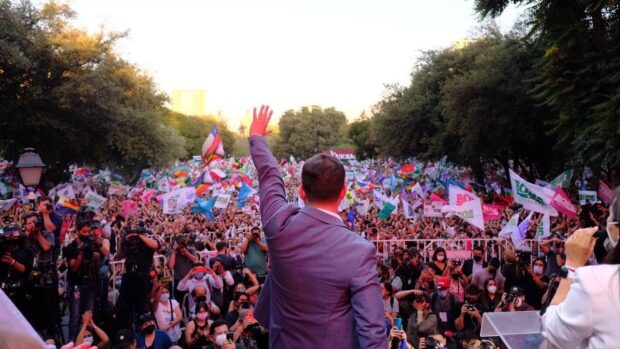 Hombre con traje saludos a una multitud de personas. En la multitud se ven distintas banderas, de Chile de la comunidad LGBTIQ+ y otras que dicen Boric.
