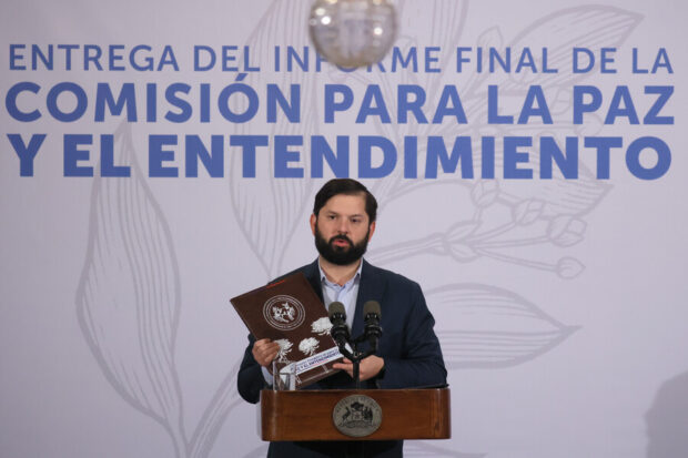 Presidente Boric da discurso desde un podio, detrás de el una gigantografía que dice: Entrega del informe final de la Comisión para la Paz y el Entendimiento.