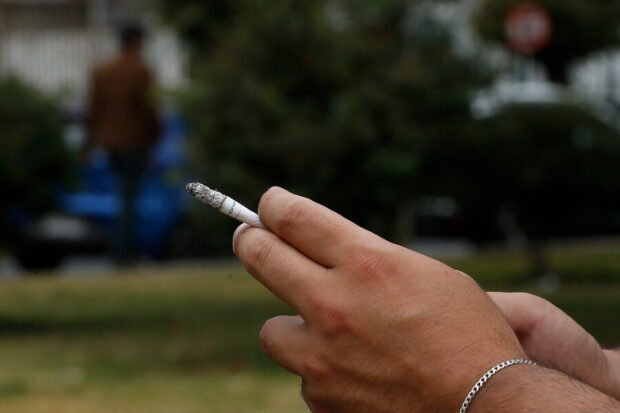 Primer plano de una mano sosteniendo un cigarro encendido, con fondo urbano desenfocado.