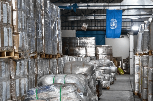 Almacenistas lleno de cajas de suministros en los estantes y en el piso arriba de palets. Del techo cuelga una bandera azul con el logo blanco de la ONU.