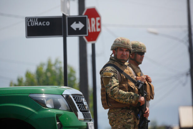 Dos militares en la calle, detrás de ellos se ve la punta de una camioneta policial y un cartel que señaliza la calle "Lumanco"