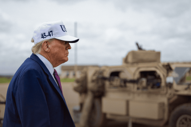 Donald Trump de perfil con una gorra blanca, al fondo se ve borrosa un tanque militar.