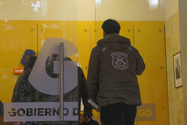 Mampara con el logo del Gobierno de Santiago, saliendo a través de la puerta se ven funcionarios de espaldas con la chaqueta verde de Carabineros.