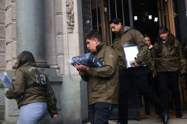Funcionarios de Carabineros con chaquetas verdes saliendo de un edificio mientras cargan carpetas y aparatos electrónicos.