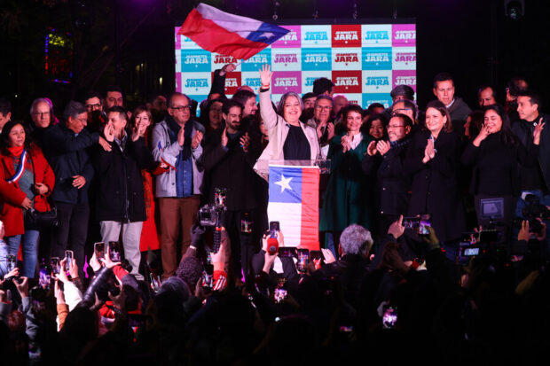 Jeannette Jara detrás de un podio con la bandera de Chile, detrás de ella figuras políticas de su sector.
