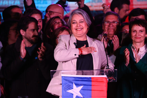 Jeannette Jara detrás de un podio con la bandera de Chile, detrás de ella figuras políticas de su sector.