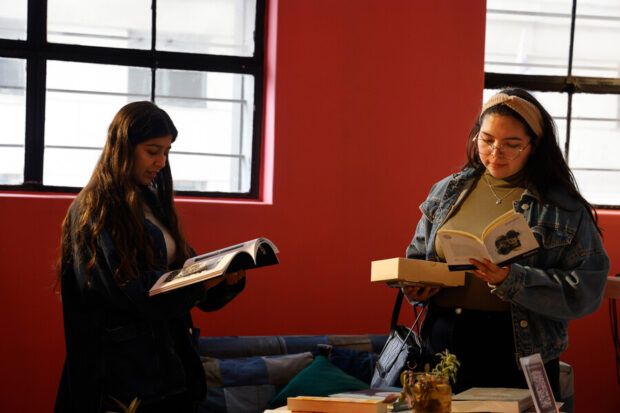 Dos mujeres jóvenes leyendo libros en una habitación con paredes rojas y ventanas grandes.
