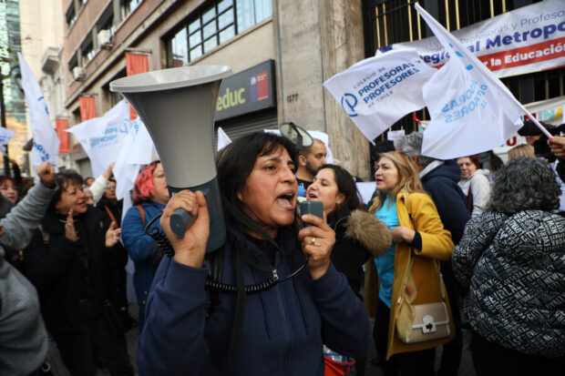 Mujer dice consignas a través de un megáfono. A su alrededor hay manifestantes, algunos de ellos con la bandera blanca y de letras azules que dice "Colegio de Profesoras y Profesores".