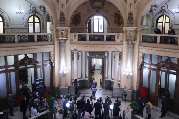 Un salon de techo alto, estructura de mármol y grandes ventanas. En el centro se ve al gobernador Orrego desde un podio mientras se dirige a los periodistas y camarógrafos que tiene al frente.
