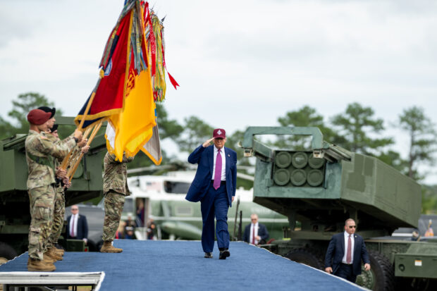 Trump desfila con un gorro rojo en una base militar. Se ve machando de frente, unos militares con banderas a su derecha y a su izquierda un tanque militar.