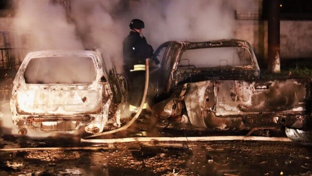 Funcionario del Servicio de Estado de Emergencia de Ucrania con la respectiva chaqueta negra y líneas reflectantes en medio de dos autor totalmente calcinados. El funcionario sujeta una manguera mientras rocía agua dentro de uno de los autos.