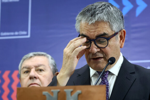 Primer plano del Ministro de Hacienda de Chile, Mario Marcel, con gafas, tocándose la frente mientras habla ante un micrófono, con otro hombre en el fondo.