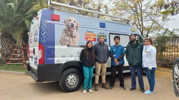Ambulancia veterinaria con una cara de un perro estampada. Al lado del vehículo, cinco médicos veterinarios con uniformes clínicos