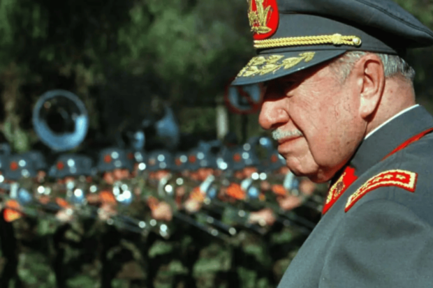 Augusto Pinochet fotografiado de perfil, viste traje militar y al fondo se ven militares en fila.