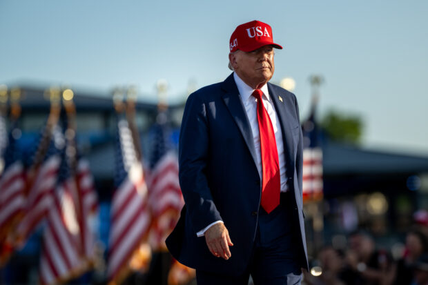 Trump caminado mientras usa un jockey rojo con las siglas USA. Detrás de él se ven banderas de EE.UU.