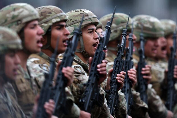Grupo de soldados en formación, vestidos con uniforme de camuflaje y portando fusiles, marchan al unísono durante un desfile militar.