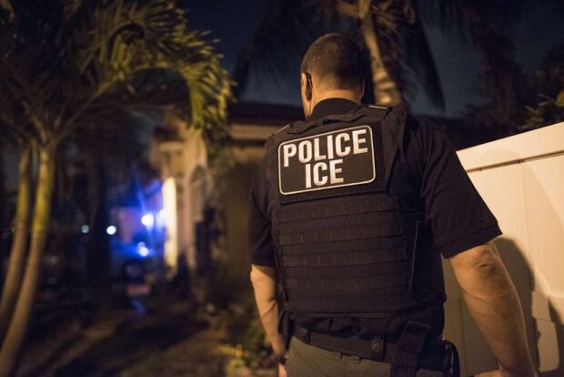 Agente de ICE con chaleco táctico patrullando de noche frente a una residencia..