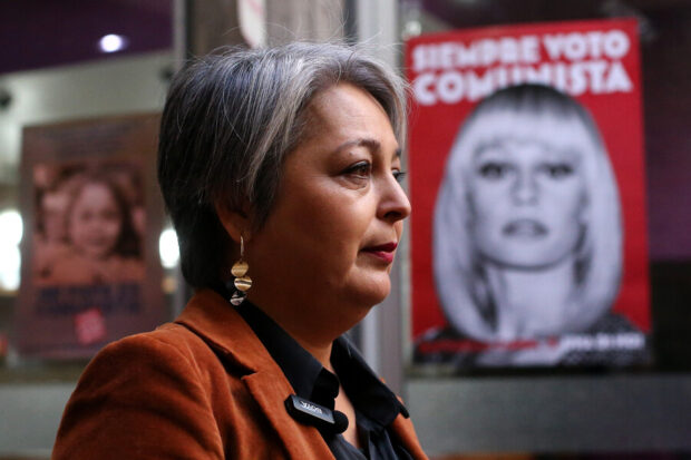 Jeannette Jara es fotografiada de perfil. Detrás de ella se ve un cartel con la imagen de una mujer y que dice: "Siempre voto comunista".
