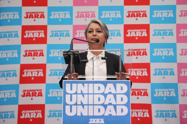 Jeanette Jara se dirige al público desde un podio con un cartel que prominentemente muestra la palabra 'UNIDAD' y su nombre. El fondo está decorado con repetidos logos de su campaña.