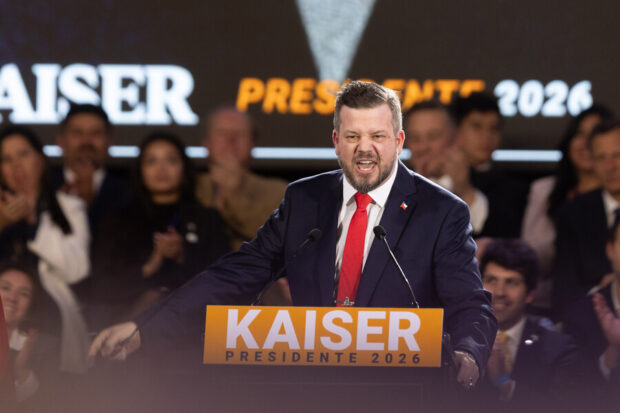 Johannes Kaiser desde un podio que dice KAISER con un fondo naranja. Detrás de él adherentes.