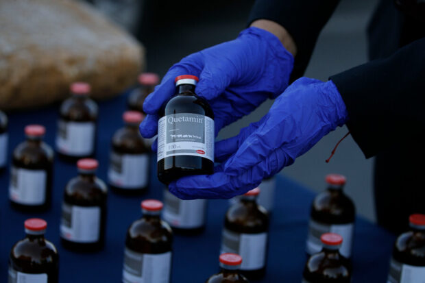 Manos con guantes azules sostienen una botella, en su etiqueta se lee "ketamina".
