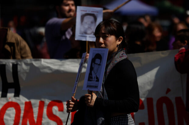 Mujer sostiene una imagen de una persona detenida desaparecida que tiene la pregunta "¿Dónde están?". Detrás de ella manifestantes con más imágenes en blanco y negro de víctimas.