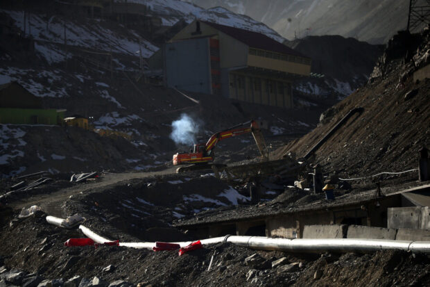 Excavadora trabajando en un terreno rocoso de la mina El Teniente, con una columna de humo saliendo del motor, rodeada de estructuras industriales y montañas parcialmente cubiertas de nieve.