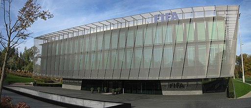 Fachada moderna del edificio principal de la sede de la FIFA en Zúrich, Suiza, con una estructura de vidrio y metal en tonos grises, rodeada de áreas verdes.