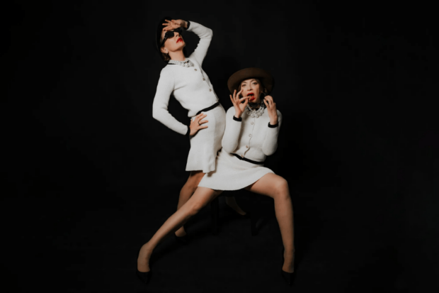 Antonia Zegers y Patricia Rivadeneira calzando trajes blancos y sombreros, con lentes de sol y zapatos negros, en una de las escenas de la obra "Una mujer llena de vicios"