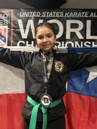 Primer plano de Camila Pérez, la joven bicampeona mundial de Kung Fu, con su uniforme, cinturón verde, medalla y la bandera chilena detrás.