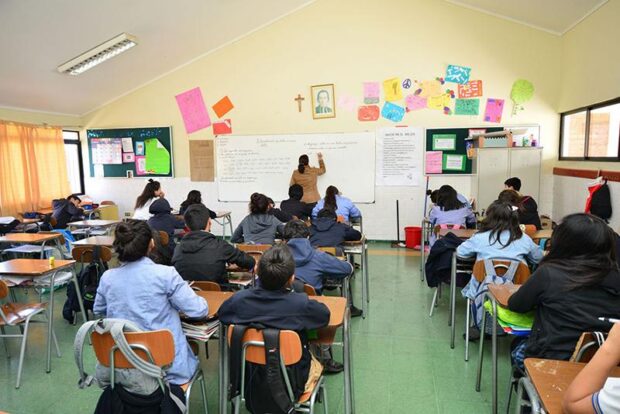 Una profesora escribe en el pizarrón blanco mientras los alumnos están sentados en los pupitres del aula escolar.