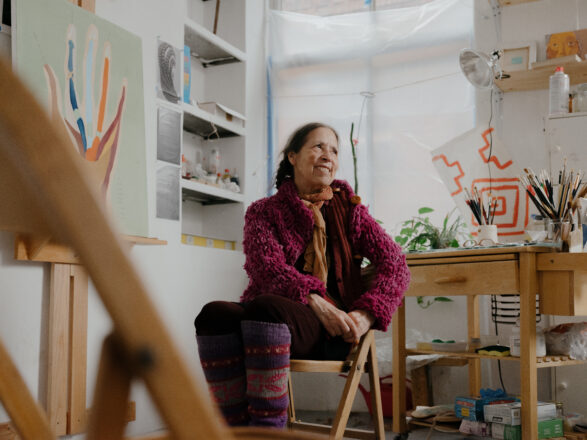 Cecilia Vicuña sentada en su taller, rodeada de pinturas, pinceles y materiales de arte.