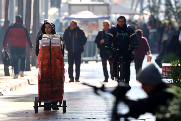 Un joven trabajador con capucha empuja un carro de mano cargado de periódicos envueltos en plástico rojo, mientras transita por una calle soleada y concurrida de Santiago, Chile, con transeúntes y edificios borrosos de fondo.