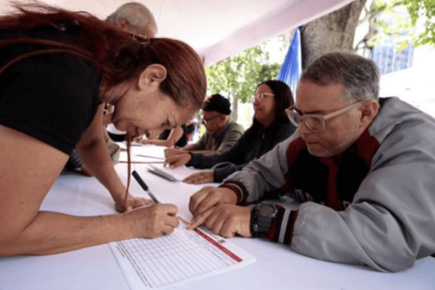 Dos adultos, una mujer y un hombre, están sentados en una mesa al aire libre con otras personas. La mujer, que tiene el cabello rojizo, se inclina sobre la mesa para llenar un formulario. El hombre que está a su lado, con cabello gris y gafas, la ayuda señalando una fila en el documento.