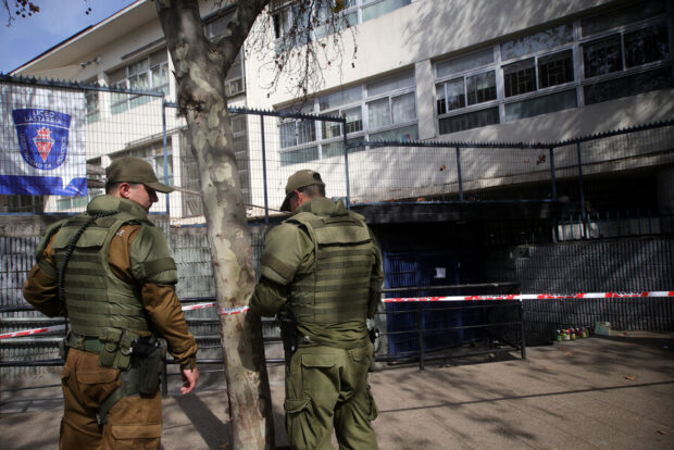 Dos carabineros con equipo táctico de espaldas, resguardando la entrada de un liceo. La imagen muestra un cordón de seguridad de cinta y botellas en el suelo, indicando que hubo incidentes recientes. Al fondo, se ve la fachada del Liceo José Victorino Lastarria y una bandera con el escudo de la institución