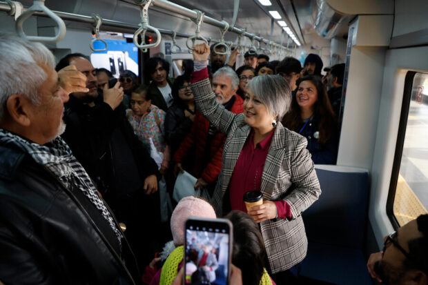 Jeannette Jara se sujeta de una agarradera tipo argolla que cuelga del techo del vagón del metro. La candidata se ve rodeada de personas mientras sonríe.