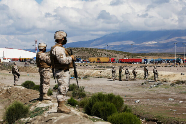 Fotografía de militares chilenos en primer plano, armados y equipados, vigilando un campamento de migrantes en una zona fronteriza árida. Al fondo se aprecian montañas, vehículos de carga y un grupo numeroso de personas, lo que sugiere una situación de control migratorio en el desierto del norte de Chile.
