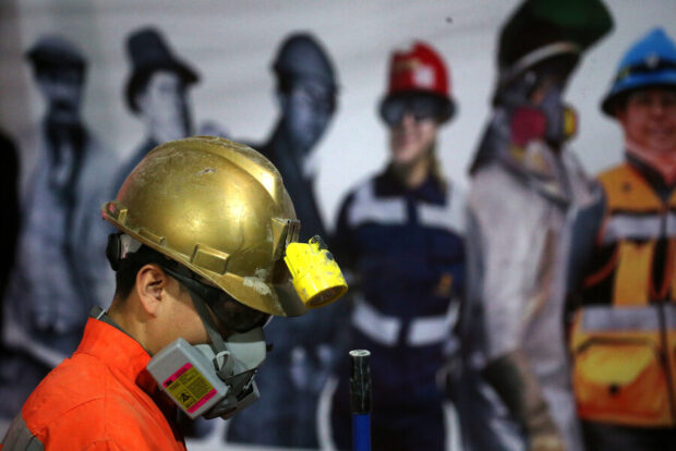 Primer plano de un minero de El Teniente con casco de oro y mascarilla, con siluetas borrosas de otros trabajadores de fondo.