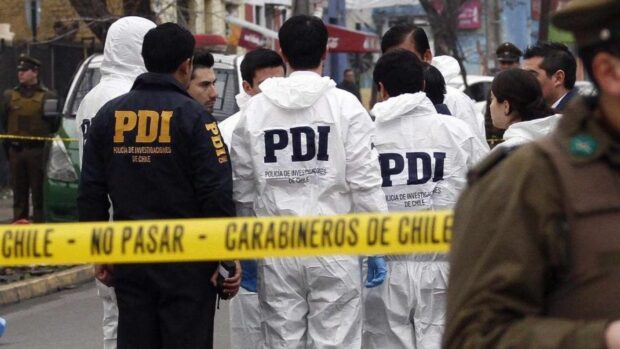 Un grupo de detectives de la PDI de Chile trabajando en un lugar de investigación. Varios de los oficiales llevan overoles blancos. En primer plano, una cinta de seguridad amarilla con los logos de la PDI y Carabineros de Chile restringe el acceso.