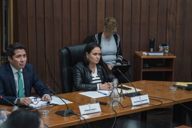 Reunión formal en mesa de trabajo con tres personas visibles, una mujer al centro con chaqueta de cuero negra y un hombre con traje azul y corbata verde, en una sala con paredes de madera.