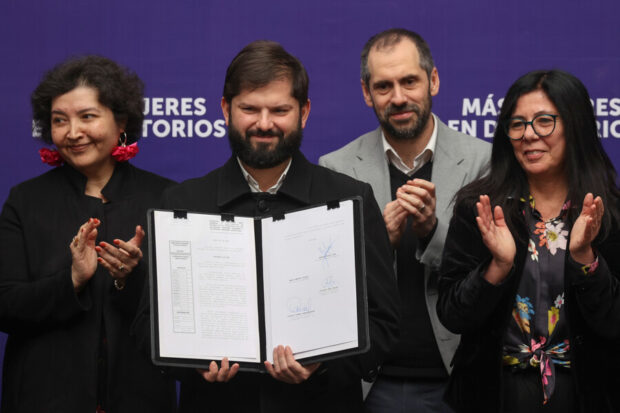 El Presidente Gabriel Boric, barbudo y vestido con abrigo negro, sostiene un documento abierto con firmas. Lo flanquean la ministra Claudia Donaire y la subsecretaria Javiera Petersen, ambas aplaudiendo. Detrás, el ministro Nicolás Grau también aplaude.