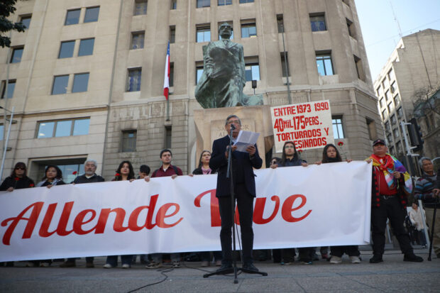 Lautaro Carmona entrega discurso de pie por un micrófono. Detrás de él, militantes del PC sostienen una pancarta blanca y con letras rojas que dice "Allende vive".
